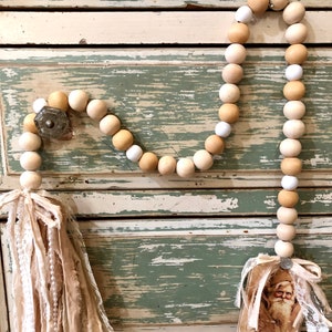 May include: A wooden bead garland featuring white and beige beads, a fabric tassel, and a vintage Santa Claus image attached. The garland is displayed on a distressed wooden surface.