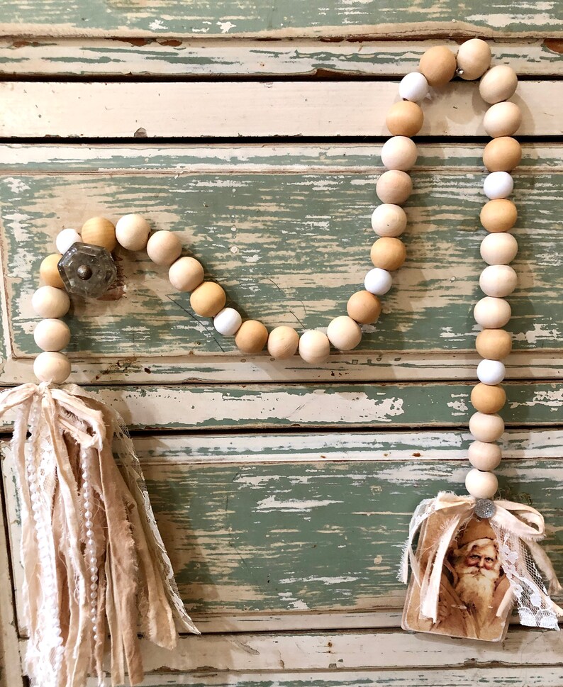 May include: A wooden bead garland featuring white and beige beads, a fabric tassel, and a vintage Santa Claus image attached. The garland is displayed on a distressed wooden surface.
