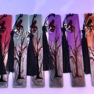 May include: A set of ten black and silver skull and crossbones bookmarks with black tassels. The bookmarks are made of resin and have a variety of colors, including red, purple, green, and brown. Each bookmark features a silhouette of a raven and a bare tree.