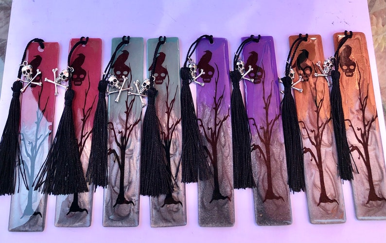 May include: A set of ten black and silver skull and crossbones bookmarks with black tassels. The bookmarks are made of resin and have a variety of colors, including red, purple, green, and brown. Each bookmark features a silhouette of a raven and a bare tree.