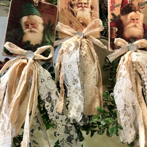 May include: Three vintage Christmas card ornaments with images of Santa Claus. Each ornament features a white lace trim and a beige fabric bow with a silver embellishment.