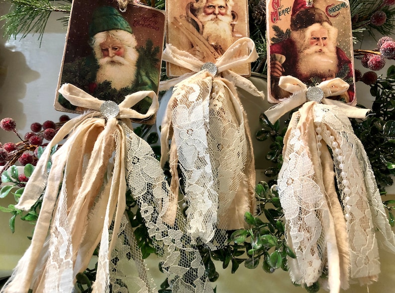 May include: Three vintage Christmas card ornaments with images of Santa Claus. Each ornament features a white lace trim and a beige fabric bow with a silver embellishment.