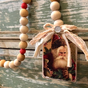 May include: A wooden bead garland with red and beige beads hangs on a light green and white wood panel. A vintage Santa Claus card with a white ribbon is attached to the garland.