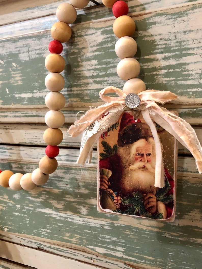 May include: A wooden bead garland with red and beige beads hangs on a light green and white wood panel. A vintage Santa Claus card with a white ribbon is attached to the garland.