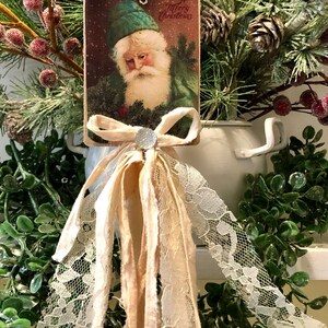 May include: Green Santa Christmas ornament with a vintage postcard image of Santa Claus wearing a green hat and a white beard. The ornament is decorated with a white lace trim and a beige ribbon bow with a silver rhinestone.