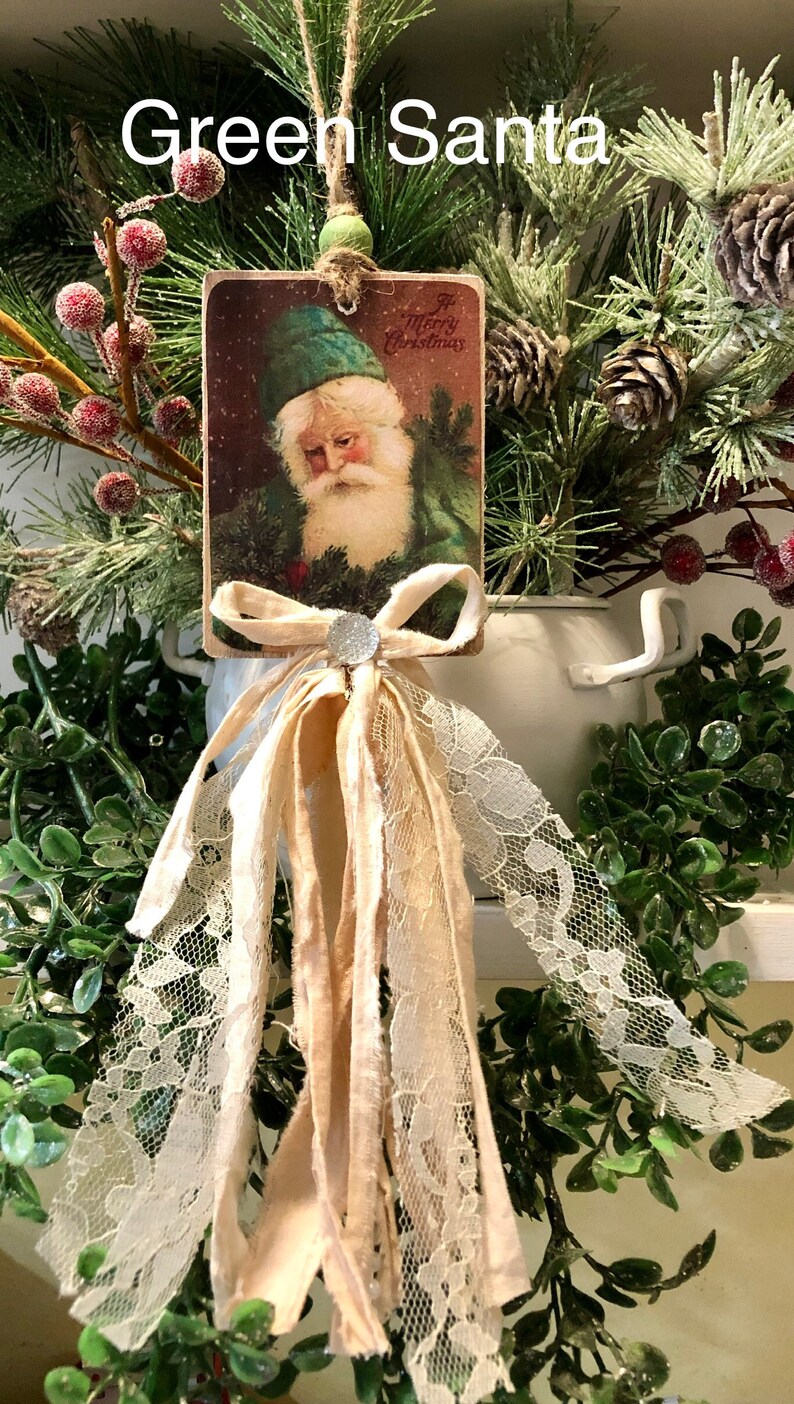 May include: Green Santa Christmas ornament with a vintage postcard image of Santa Claus wearing a green hat and a white beard. The ornament is decorated with a white lace trim and a beige ribbon bow with a silver rhinestone.