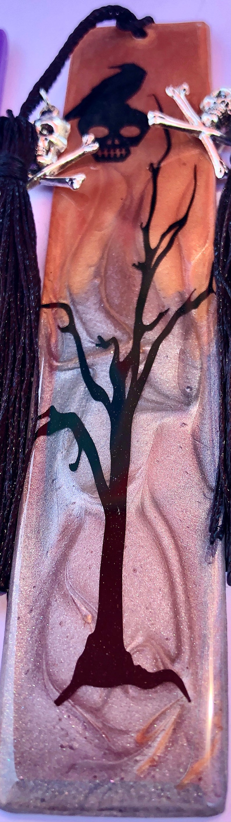 May include: A brown and silver resin bookmark with a black silhouette of a tree and a skull with crossbones. The bookmark has a black tassel.