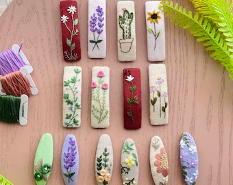 May include: A set of 14 handmade hair clips featuring embroidered floral designs. The clips are made of fabric and come in a variety of colors, including green, purple, yellow, red, and white. The designs include flowers, leaves, and a cactus.