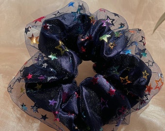 May include: A navy blue hair scrunchie with a sheer mesh overlay. The mesh is decorated with colorful glitter stars.