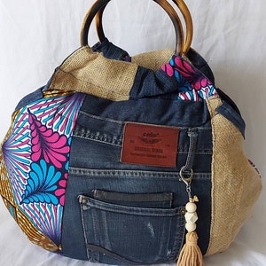 Può includere: Una grande borsa tote patchwork marrone, blu e rosa con manici in legno. La borsa è realizzata in denim e tessuto riciclati. Ha una piccola tasca sul davanti e una toppa in pelle marrone con il testo "Callio Original Denim" su di essa. Un portachiavi con perline di legno e nappa pende dalla borsa.