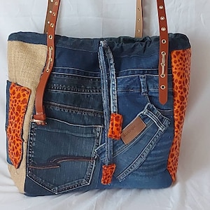 Può includere: Una grande borsa tote realizzata in tessuto denim riciclato. La borsa ha una tracolla in pelle marrone e un design patchwork con accenti in tessuto arancione e marrone. La borsa ha una grande tasca sul davanti e una tasca più piccola sul lato.