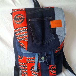 Puede incluir: Una mochila vaquera con detalles en rojo y negro. La mochila tiene cierre de solapa, múltiples bolsillos y correas ajustables. El diseño incorpora una etiqueta con el texto "OBAMSBAGS".