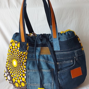 Peut inclure: Un sac cabas en jean bleu avec un panneau de tissu à motifs jaunes et blancs. Le sac a des anses en cuir marron et une fermeture à cordon. Le sac a une poche sur le devant et une étiquette qui dit "DENIM SOUL COLLECTION".
