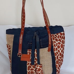 Peut inclure: Un sac cabas brun et beige en denim recyclé et en toile de jute avec un panneau en tissu imprimé girafe. Le sac est doté de sangles en cuir brun avec des œillets dorés et une petite étiquette en cuir brun avec le texte "OBAMBAGS".