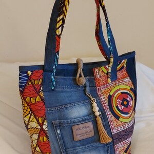 Peut inclure: Un sac cabas en jean bleu avec des panneaux en tissu imprimé africain coloré. Le sac a une grande poche sur le devant avec le texte "PULL&BEAR" et un pompon brun suspendu à la tirette de la fermeture éclair.