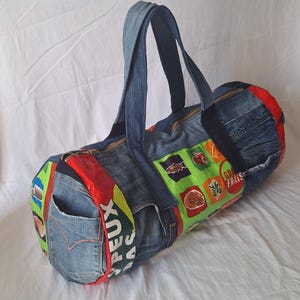 Può includere: Una borsa da viaggio in denim con un design patchwork. La borsa presenta una chiusura con cerniera, manici in denim e una tasca. Il patchwork include accenti rossi, verdi e gialli, con dettagli di testo e grafici. Realizzata in denim riciclato.