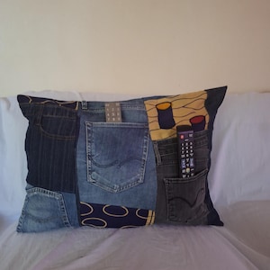 Könnte beinhalten: Ein rechteckiges Kissen aus recycelten Jeans. Das Kissen zeigt verschiedene Blautöne von Denim-Flicken, mit sichtbaren Taschen. Eine Fernbedienung befindet sich in einer der Taschen. Das Kissen liegt auf einer weißen Oberfläche.