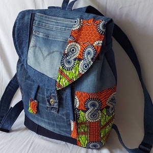 Könnte beinhalten: Ein Jeans-Rucksack mit Klappenverschluss, Taschen und Riemen. Die Tasche ist mit leuchtend orangefarbenem, grünem und blau gemustertem Stoff akzentuiert. Der Rucksack hat einen Griff und verstellbare Schultergurte.