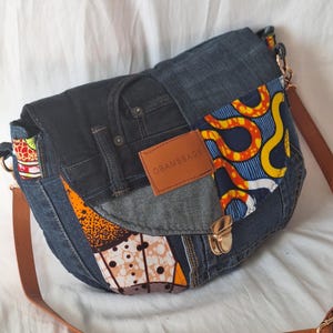 Op de afbeelding: Een denim crossbody tas met een klepsluiting. De tas heeft een patchwork denim constructie met kleurrijke stoffen accenten. Het heeft een bruine leren riem en een goudkleurige sluiting. Het label op de tas luidt "OBAMSBAGS".