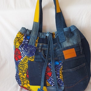 Peut inclure: Un sac cabas en jean bleu avec des anses jaunes et une fermeture à cordon. Le sac est fabriqué à partir de denim recyclé et présente un panneau en tissu imprimé africain coloré. Le sac porte une étiquette en cuir marron qui dit "OBAMSAGS".