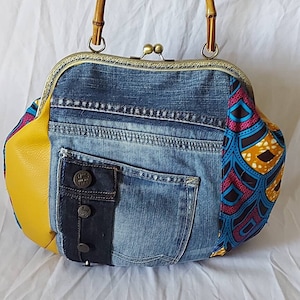 Può includere: Una borsa a mano in denim blu con un pannello in pelle gialla, una tasca in denim nero e un pannello in tessuto con motivi colorati. La borsa ha un manico in bambù e una chiusura in ottone.