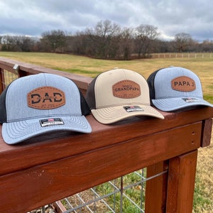 Peut inclure: Trois casquettes de baseball avec des filets noirs à l'arrière et des patchs en cuir. Les casquettes sont grises, beiges et bleues. Les patchs portent les mots "DAD EST. 2021", "Grandpa" et "Papa J".