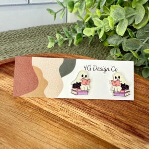 May include: A pair of ghost-shaped earrings, each with a black cat reading a book. The earrings are on a white card with the text "4G Design Co" and a colorful abstract design. The earrings are on a wooden tray.