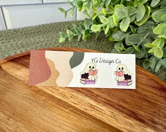 May include: A pair of ghost-shaped earrings, each with a black cat reading a book. The earrings are on a white card with the text "4G Design Co" and a colorful abstract design. The earrings are on a wooden tray.