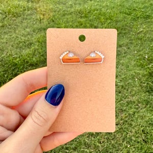 May include: A pair of earrings shaped like slices of pumpkin pie with cream. The earrings are on a brown card. The background is green. The earrings are made of a shiny material.
