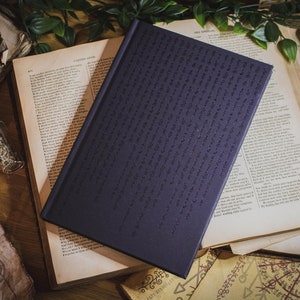 May include: A dark purple hardback journal with a cover design of a repeating pattern of small symbols. The journal is lying on top of several old, open books with yellowed pages.