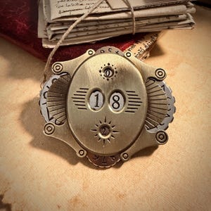 May include: A brass-toned, steampunk-inspired dial showing the number "18". The oval centre features a sunburst design and horizontal lines. The dial is framed by decorative metalwork and small circular accents. The image also shows a stack of old papers.