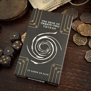 May include: A black card deck with gold detailing and the text "THE DECK OF TWENTY-TWO THINGS". The deck features a white spiral design and is surrounded by black dice with gold dots and antique coins on a dark wooden surface. The text "22 CARDS OF FATE" is also visible.