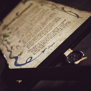 May include: An aged parchment document with ornate blue and gold border details, displayed in a black frame. A black wax seal with a starburst design rests near the document. The word "Signed" is visible.