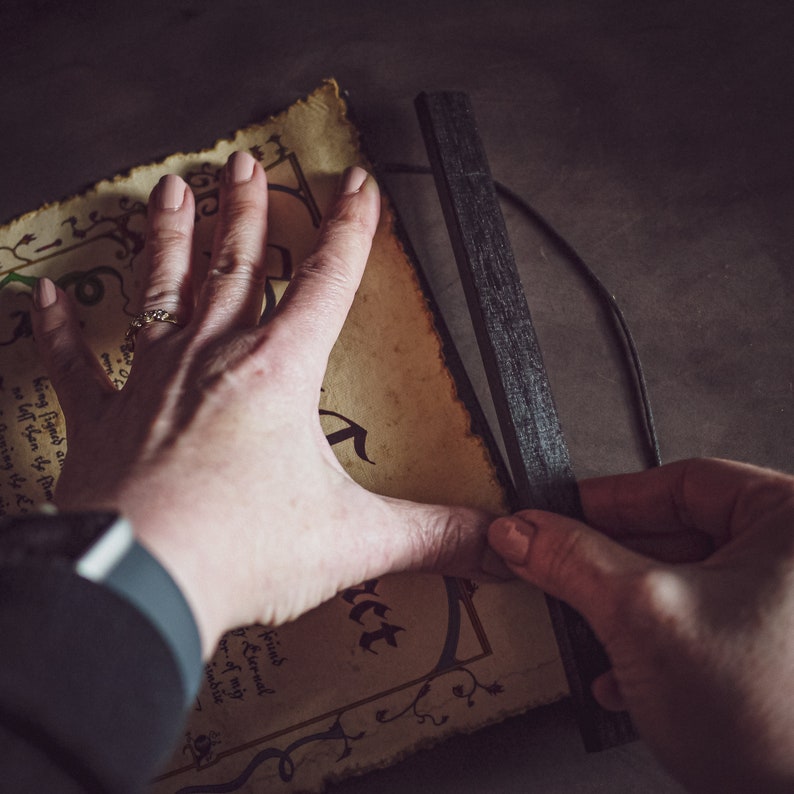May include: Aged parchment-style paper with handwritten text and decorative borders is held in place by a black wooden magnetic poster hanger. The paper has a distressed, vintage look, with visible wear and tear.
