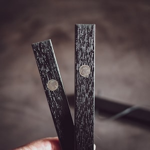 May include: Close-up of two black wooden magnetic poster hangers. Each hanger has a silver-colored magnet. The wood grain is visible. The hangers are held in a hand.