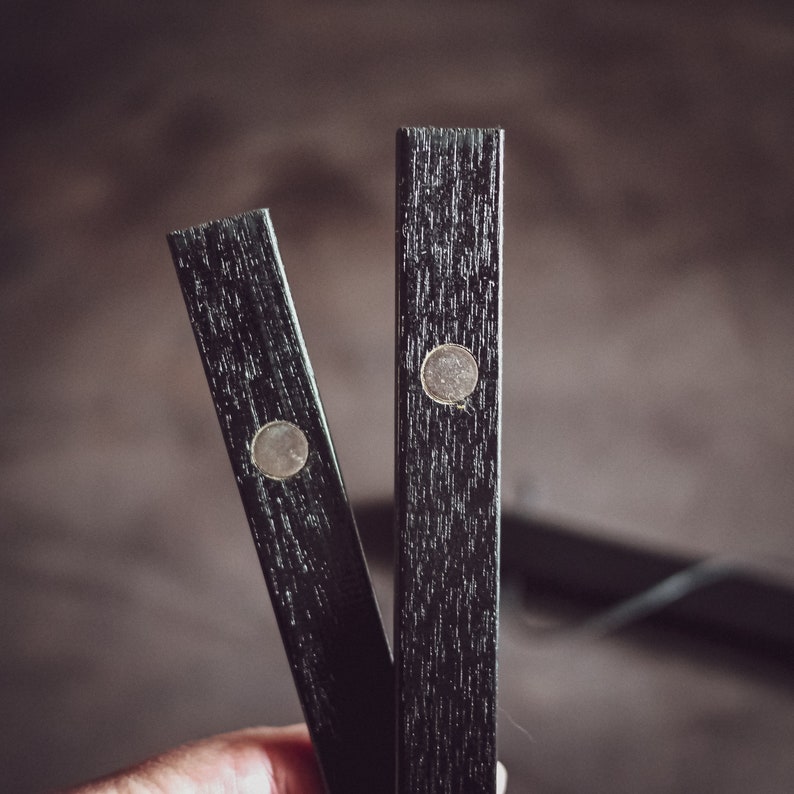 May include: Close-up of two black wooden magnetic poster hangers. Each hanger has a silver-colored magnet. The wood grain is visible. The hangers are held in a hand.