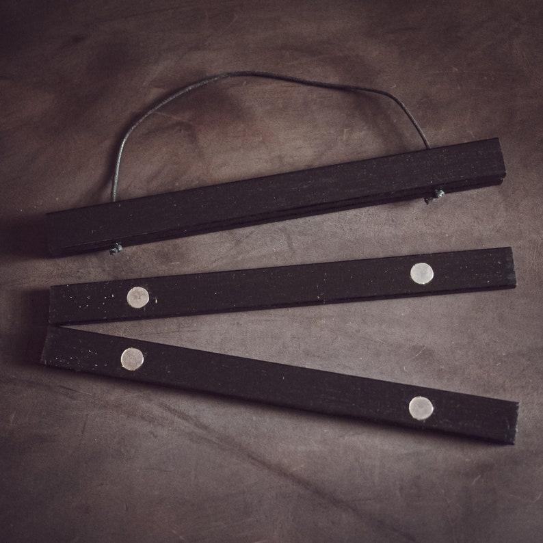 May include: Black wooden poster hanger with a leather cord for hanging. The hanger is made of three separate pieces of wood, with small magnets to hold the poster in place. The wood has a matte finish.