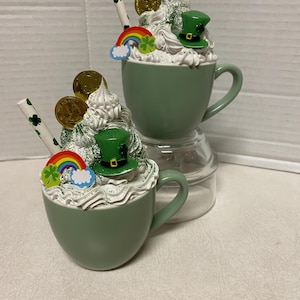 Peut inclure: Deux tasses en c&eacute;ramique vertes avec de la cr&egrave;me fouett&eacute;e blanche, des pi&egrave;ces d'or, un tr&egrave;fle vert, un arc-en-ciel et un chapeau de lutin vert. Les tasses sont sur un socle en verre transparent.