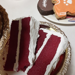 May include: Two slices of red velvet cake with white frosting on a silver platter. The cake has a deep red color and a textured appearance. In the background, there are decorated cookies and a candle.