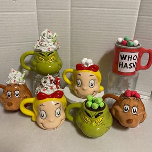 May include: Set of six Grinch and Cindy Lou Who themed ceramic mugs. Each mug features a different character from the popular holiday movie. The mugs are decorated with festive colors and designs, including red, green, white, and brown. The mugs are filled with marshmallows and candy.