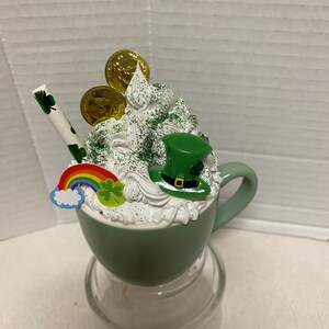 Peut inclure: Une tasse en c&eacute;ramique verte avec un bord et une poign&eacute;e blancs, remplie de cr&egrave;me fouett&eacute;e blanche, de paillettes vertes, de pi&egrave;ces d'or, d'un chapeau de l&egrave;vreau vert, d'un arc-en-ciel et d'un tr&egrave;fle. La tasse est pos&eacute;e sur un socle en verre transparent.