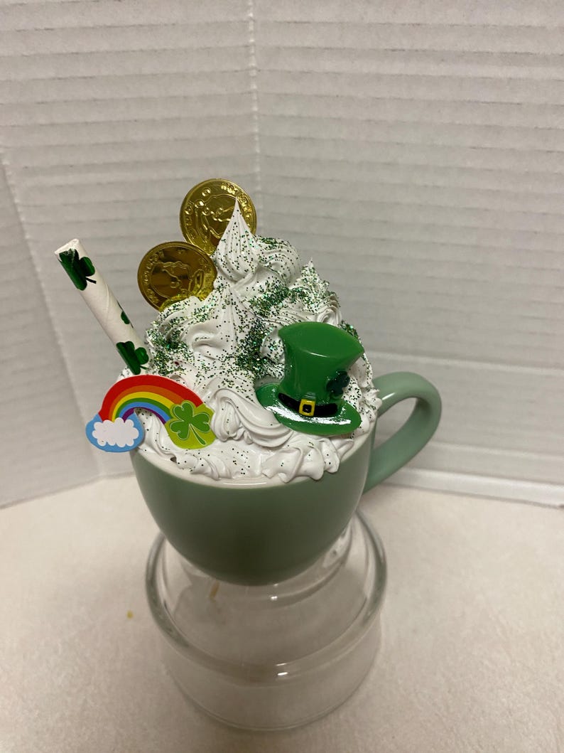Peut inclure: Une tasse en c&eacute;ramique verte avec un bord et une poign&eacute;e blancs, remplie de cr&egrave;me fouett&eacute;e blanche, de paillettes vertes, de pi&egrave;ces d'or, d'un chapeau de l&egrave;vreau vert, d'un arc-en-ciel et d'un tr&egrave;fle. La tasse est pos&eacute;e sur un socle en verre transparent.