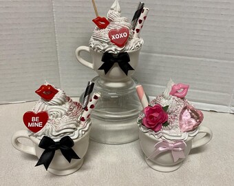 May include: Three white ceramic mugs decorated with whipped cream, candy hearts, and other Valentine's Day themed decorations. The mugs are arranged on a white surface.