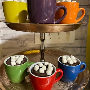 May include: A tiered serving stand with six small mugs filled with hot chocolate and marshmallows. The mugs are in various colors, including purple, orange, yellow, green, red, and blue.