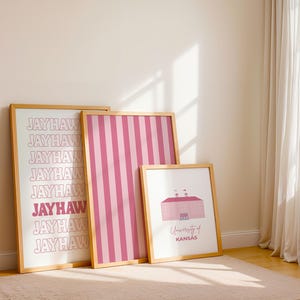 Op de afbeelding: Drie ingelijste prints leunen tegen een muur. De grootste print toont het woord "JAYHAW" herhaald in een gestapeld patroon. De middelste print heeft roze en witte verticale strepen. De kleinste print toont een roze gebouw en de tekst "University of Kansas".