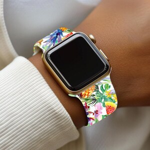 May include: A gold smartwatch with a black screen and a colourful floral band. The band features a tropical design with flowers, pineapples, and green leaves. The watch is worn on a wrist.
