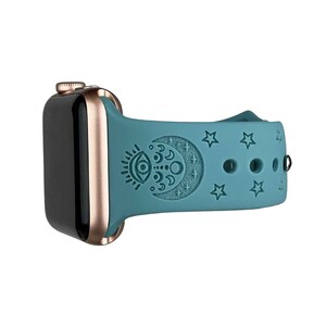 May include: A teal blue silicone watch band with a rose gold watch case. The band has a moon and eye design with stars.