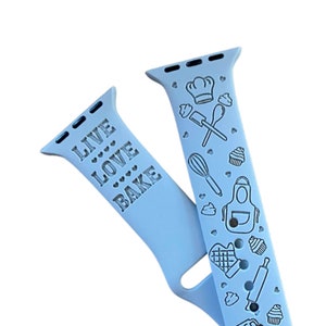 May include: A light blue silicone Apple Watch band with a white design. The band has the text "LIVE LOVE BAKE" on one side and a pattern of baking-themed icons on the other side.