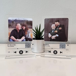May include: Two clear acrylic music player stands with photos and song titles. The first stand has a photo of a couple and the song title "61 Seconds" by "The Outfield". The second stand has a photo of two men and the song title "Songbird" by "Fleetwood Mac".