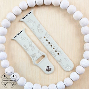 Pu&ograve; includere: Due cinturini in silicone verde chiaro per Apple Watch con stampa di cactus. I cinturini sono mostrati su una superficie in legno con perline bianche attorno al bordo.